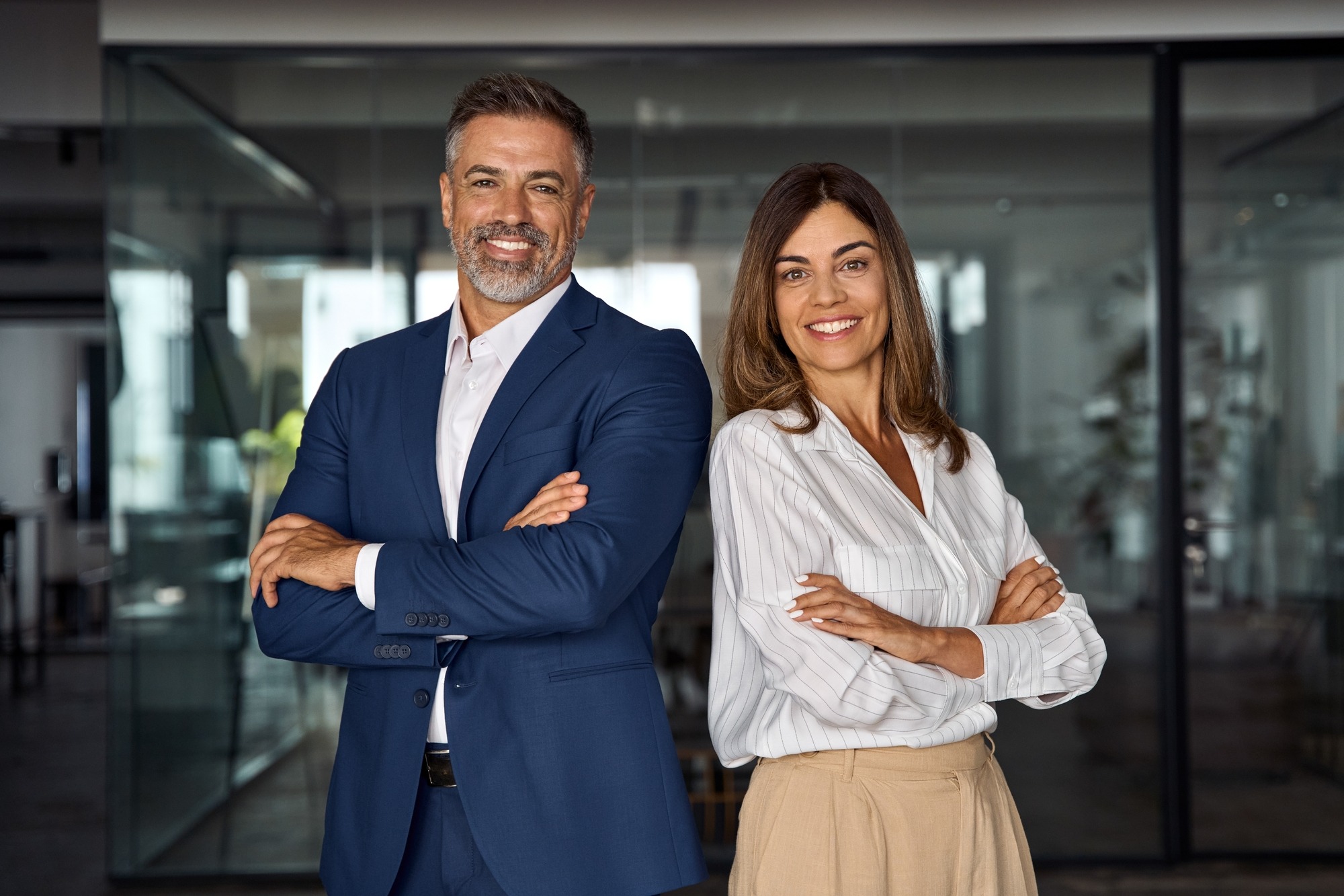 Portrait of smiling mature Latin or Indian business man and European business woman standing arms crossed in office.